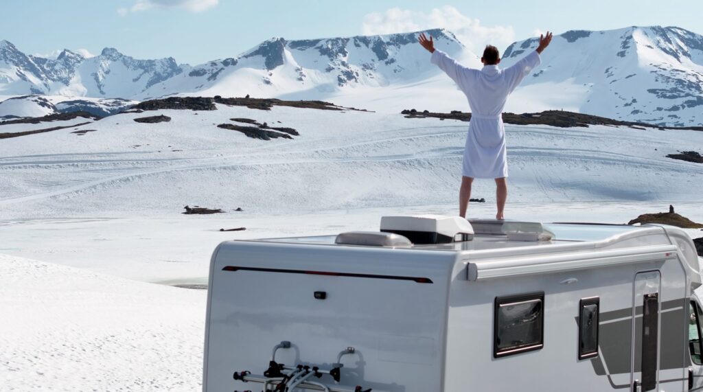 Man with his arms raised on the roof of a camper van in snowy mountains